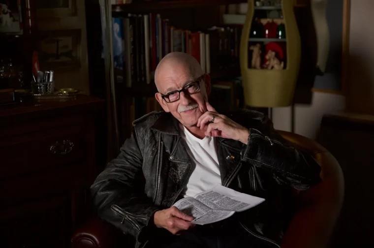 Bryan Robert Bale seated in a leather armchair holding a newspaper in his book-lined room, in a still from the documentary Bachelor, 38.