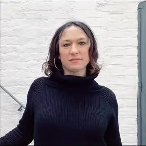 Producer Andee Ryder, with dark hair and hoop earrings, in a navy roll-neck against a white brick wall.