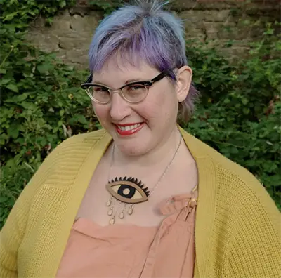 Programmer Claire Vaughan, with lilac hair and glasses, in a mustard cardigan outdoors against greenery.