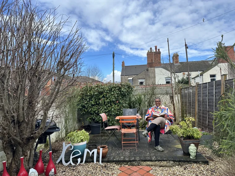 Kemi reading in her back garden, with decorative 'KEMI' letters on the ground, in the documentary Farming: Kemi's Story.