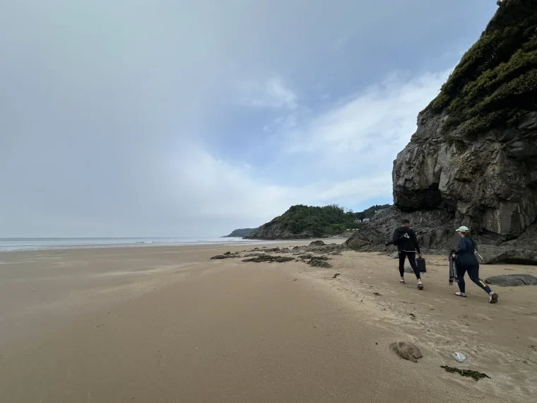 Two crew members carrying equipment across a beach beneath cliffs, behind the scenes on Everything Is Waves.