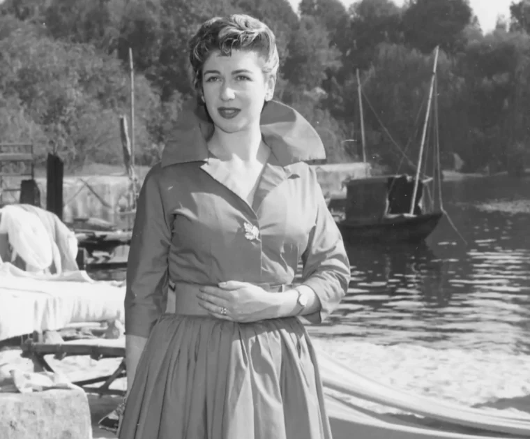 Jackie Forster as a young woman in a 1950s dress by a waterside with sailing boats, in a still from the documentary Jackie.