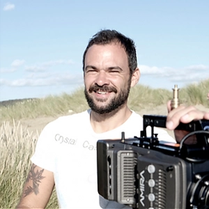 Filmmaker Jay Bedwani, a bearded man in a white T-shirt, holding a cinema camera outdoors in the dunes.